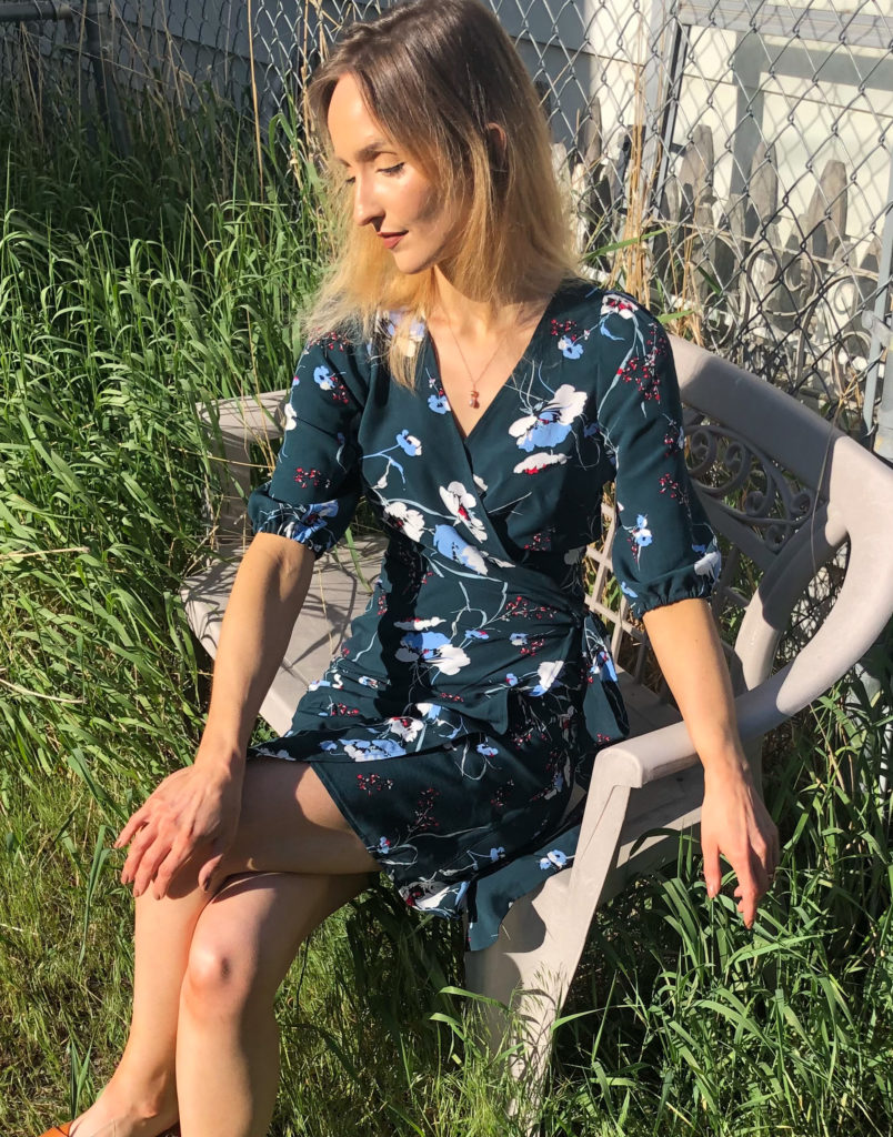 M7406 wrap dress in a teal floral print on a white woman with blonde hair sitting on a bench in tall grass. 